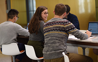 Four students work at a long table in a modern study space, with one woman smiling at a male companion while others focus on their laptops.