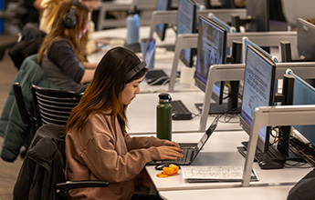 A young woman with headphones works on her laptop in a computer lab, surrounded by other students at their desks. A green water bottle and an orange snack are on her desk.