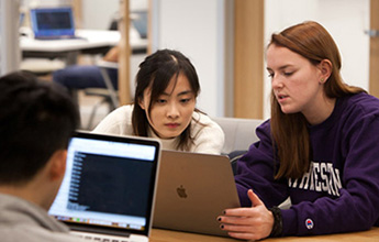 Two female students collaborate over a laptop in a study setting, while a male student works on another laptop in the background.