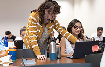 A person in a striped sweater assists a student with a laptop in a classroom.