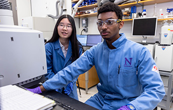 Two researchers in a lab; one seated at a computer, the other observing, both in blue lab coats and goggles.