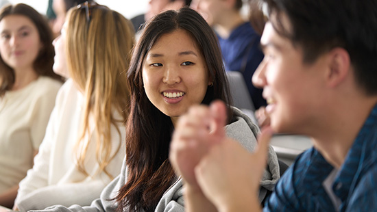Group of students sitting together in a classroom, with a smiling woman in a gray hoodie at the center and a person gesturing in the foreground.