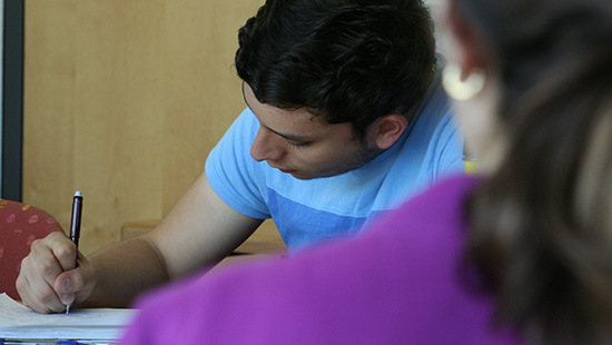 A young man writing at a desk, wearing a light blue T-shirt, with another person partially visible in the foreground.