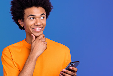 Smiling young man in an orange shirt holds a smartphone and looks thoughtfully to the right against a blue background.