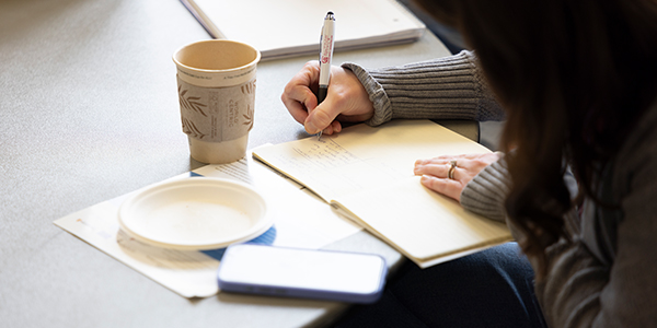 Person writing in a notepad at a table with a coffee cup and smartphone nearby.